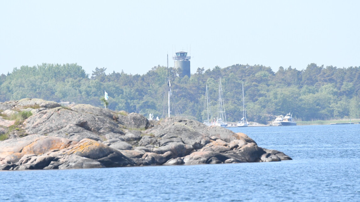 Photo taken from a boat showing blue sea in front, a rocky island behind it and furthest away, the forest like silhouette of the Örö island with a grey watchtower pointing up in the middle. 