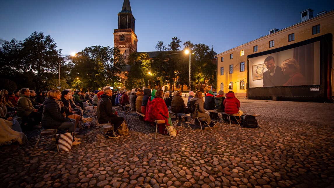 Ihmisiä ulkoilmaelokuvanäytöksessä Turun raatihuoneentorin laidalla illan hämärtyessä