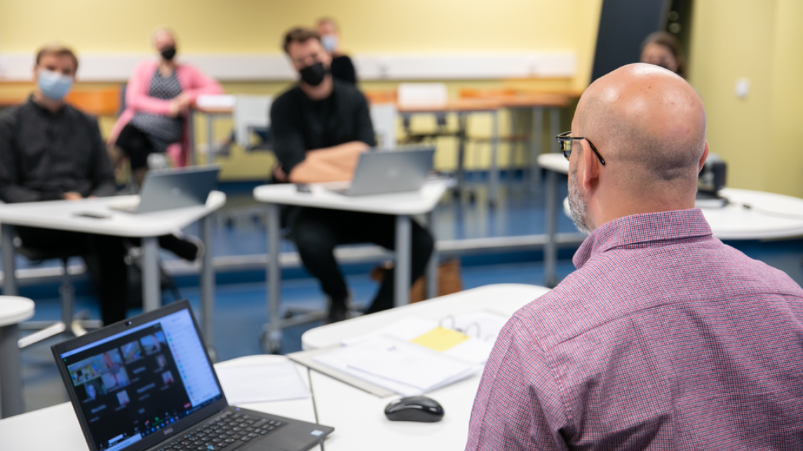 A lecturer in a hydrid teaching situation with a laptop and four students present