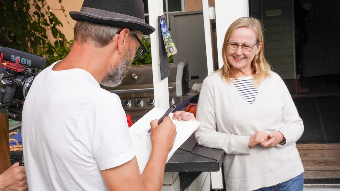 Etualalla selin kuvaajaa kohti sarjakuvapiirtäjä Ville Ranta, hattu päässä ja valkoinen t-paita yllään, pitää toisessa kädessään piirustuslehtiötä ja piirtää karikatyyriä piirtää karikatyyriä kansanedustaja Eeva-Johanna Elorannasta. Eeva-Johanna seisoo Rannan edessä, poseeraa piirtäjälle nojaten kaidetta vasten, ja nauraa iloisesti. Kuvan sivussa näkyy videokameralaitteistoa, kun toimittaja kuvaa tilannetta.