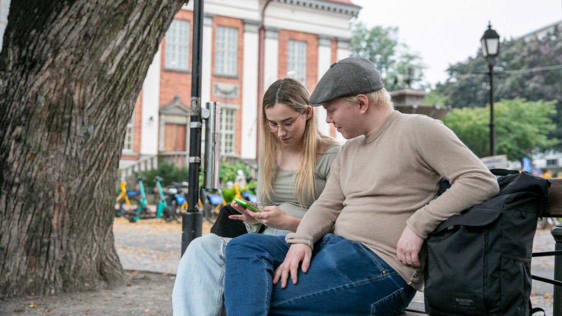 Kaksi ihmistä istuu ulkona penkillä ja katsoo puhelinta.