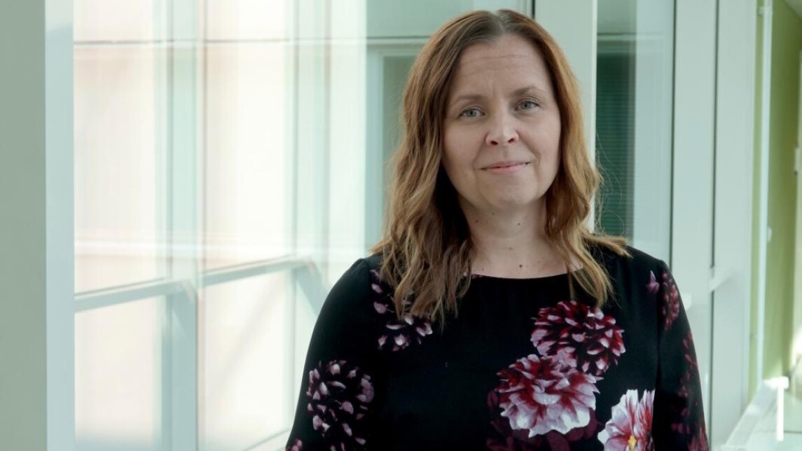 Nainen seisoo lasikäytävässä ja katsoo kameraan.