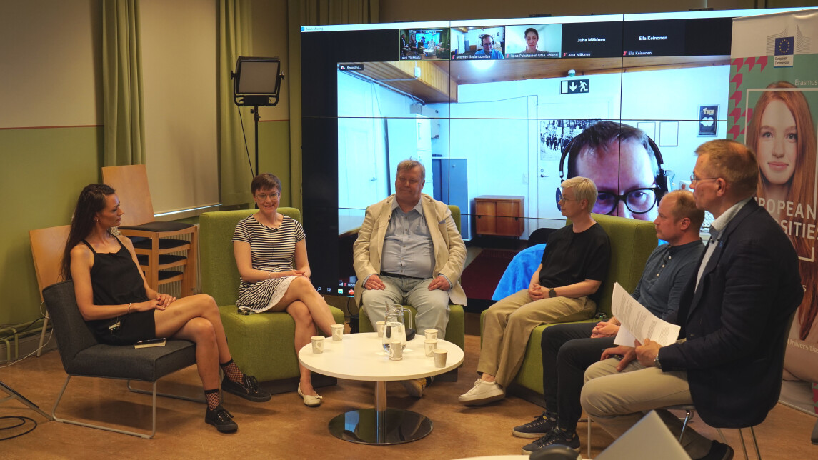 six people in a panel discussion with a screen showing remote participants in the background