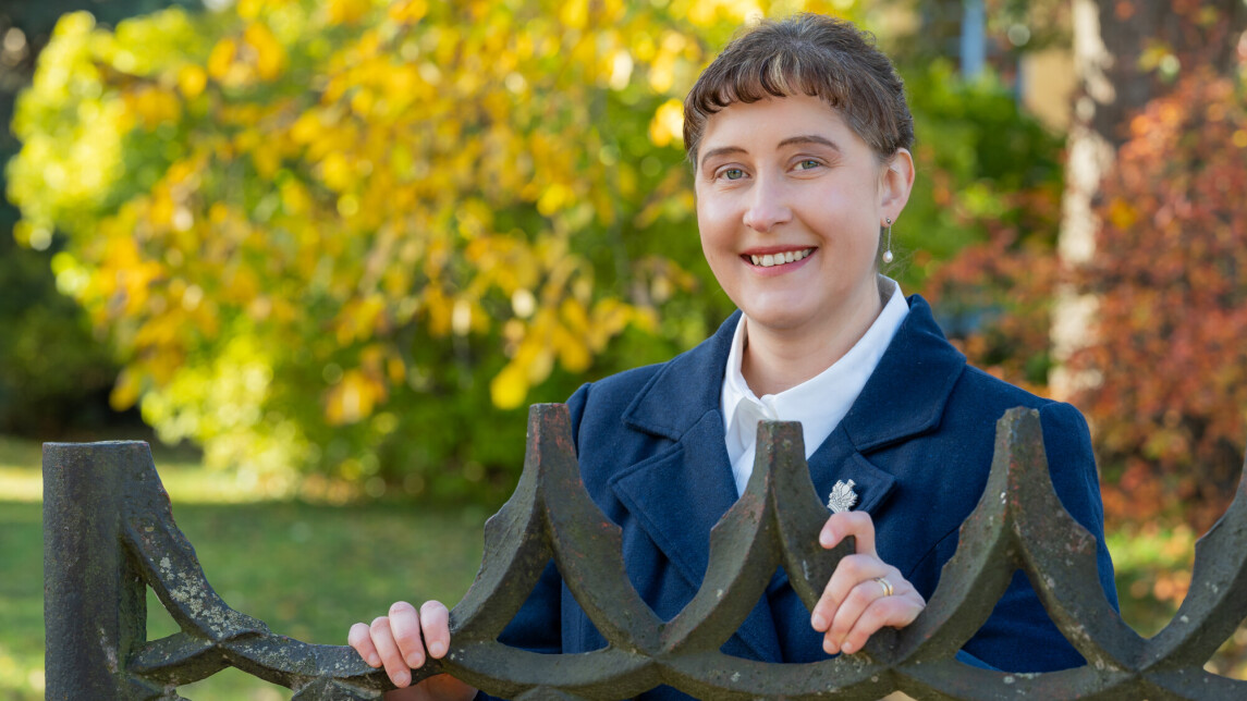 Ulla Ahlmen-Laiho lähikuvassa ulkona vanhan rautaisen portin edessä, taustalla näkyy puita joissa on värikkäät lehdet.