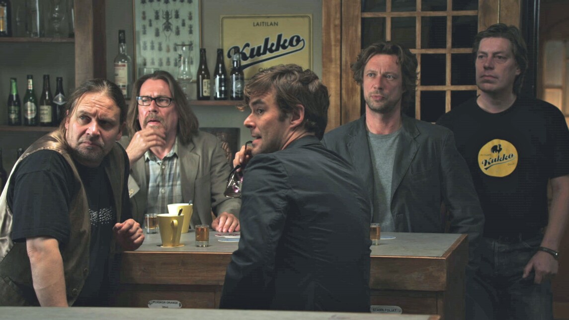 Viisi henkilöä baaritiskin molemmilla puolilla, katsovat kenties ovelle päin. 