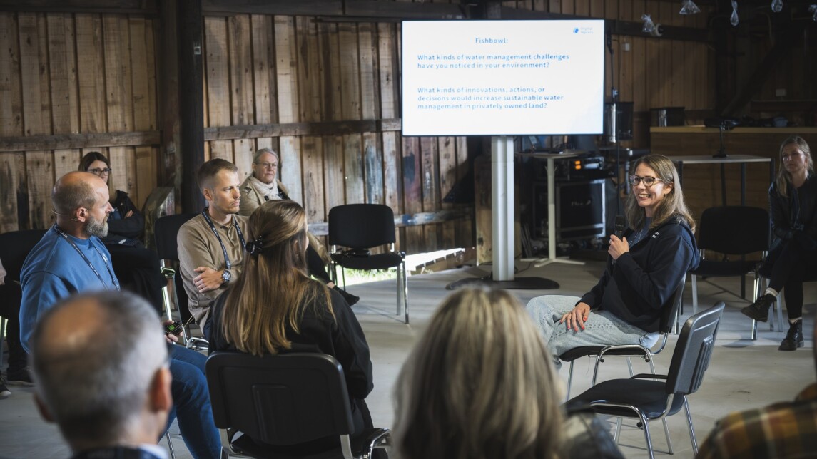 Ringissä istuvia tutkijoita keskustelemassa tilaisuudessa, kameraa kohti olevalla tutkijalla mikrofoni kädessä. Hänen takana isolla näytöllä powerpoint-esitys. Tapahtumatila saaristohenkinen, avoin iso tila jossa seinät raakalautaa ja katossa roikkuu kalastusaiheisia koristeita