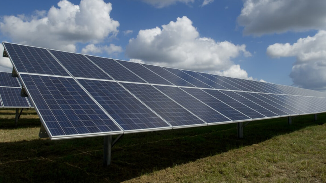 Aurinkopaneelit nurmella, taustalla sininen taivas ja pilviä.