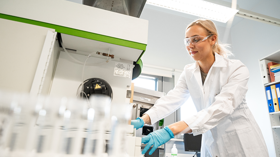 Nuori nainen työskentelee laboratoriossa yllään valkoinen labratakki, suojalasit ja kädessä siniset hanskat. Kva otettu alaviistosta ja etualalla näkyy koeputkia