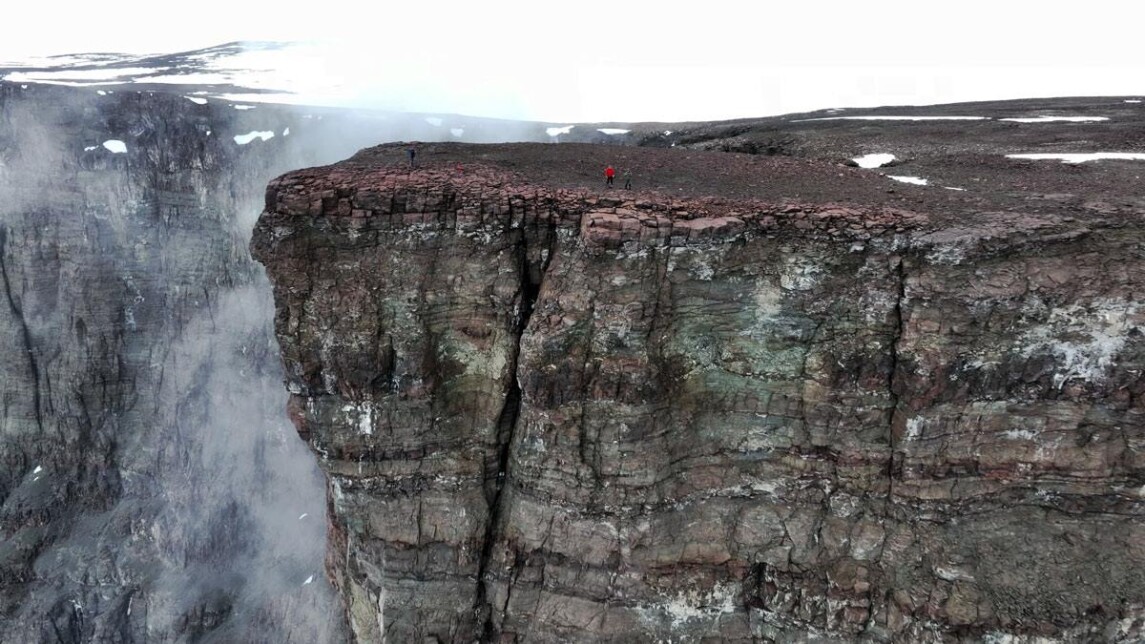 ilmakuvassa jyrkkä kallionkieleke, jonka päällä seisoo kaksi ihmistä, taustalla lunta, rotkossa nousee höyry