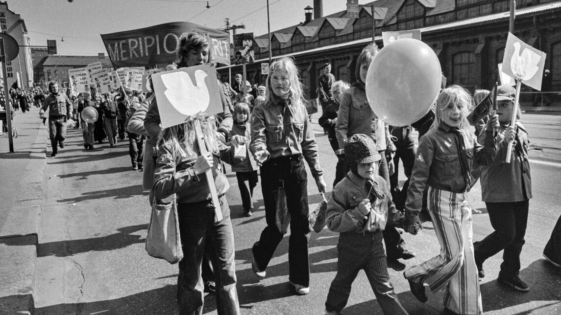 Vappumarssijoita Hämeentiellä. Edessä lapset kantavat kylttejä, joissa rauhankyyhkyn kuva. Keskellä edessä lapsi ilmapallon kanssa. Takana kannetaan banderollia, jossa teksti: Meripioneerit.