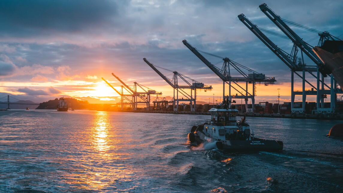 Vene ajaa kohti auringonlaskua satamassa. Taustalla isoja nostureita.