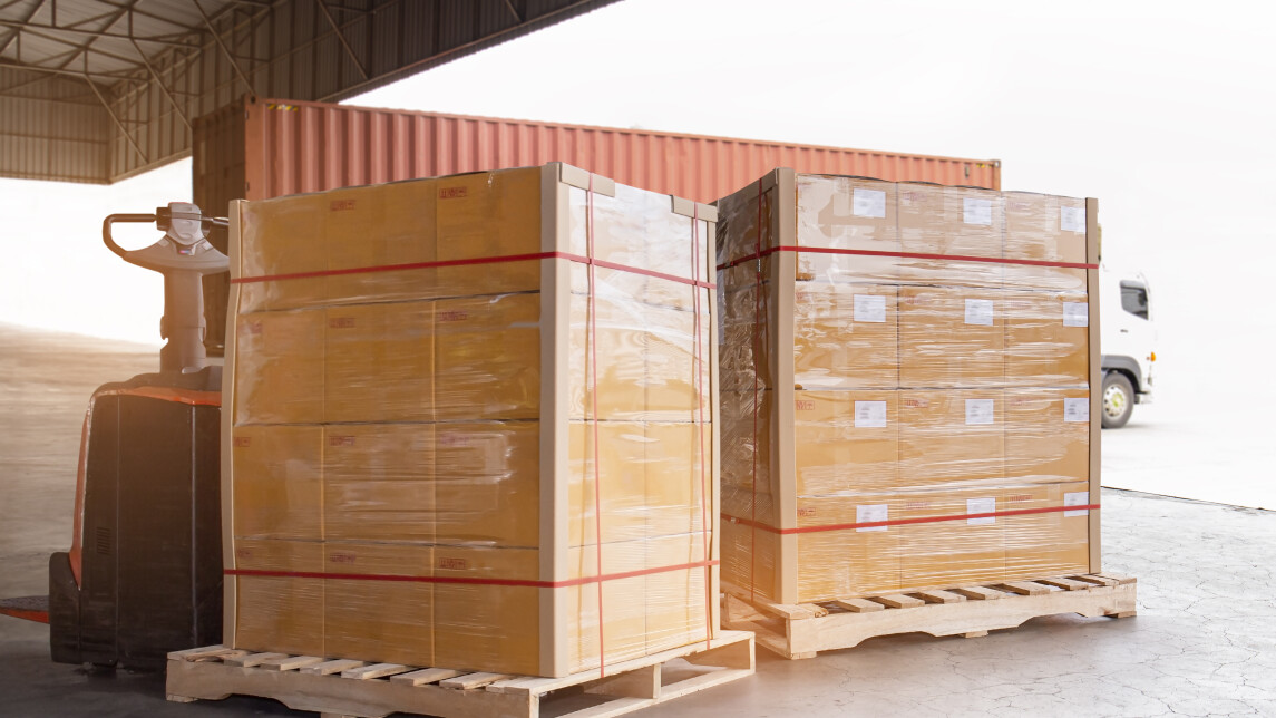 Kelmutettuja pahvilaatikoita odottaa puisilla lavoilla lastausta trukilla konttiin. Taustalla merikontti kuorma-auton kyydissä.