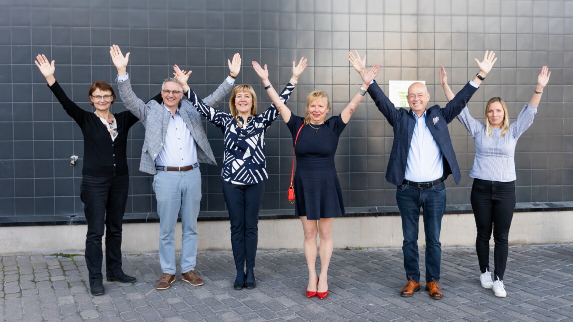 Kuusi henkilöä seisoo harmaan rakennuksen edessä kädet ilmassa.