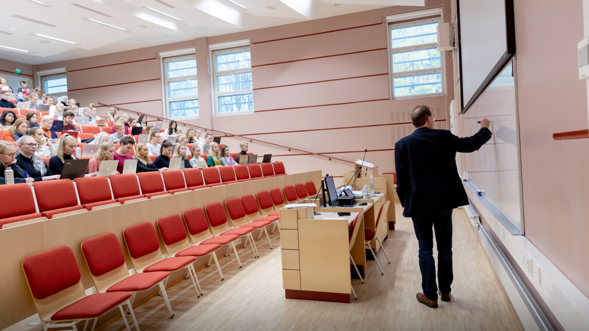 Opettaja luentosalin edessä luennoimassa suurelle joukolle opiskelijoita.