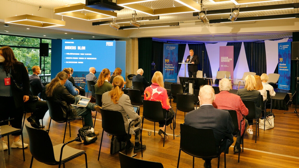 Suuri joukko turvavälein varustettuja ihmisiä kuuntelemassa luentoa.