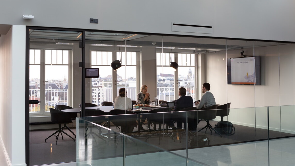Neljä ihmistä pitää kokousta pöydän ääressä tilassa, jonka seinät ovat lasia.