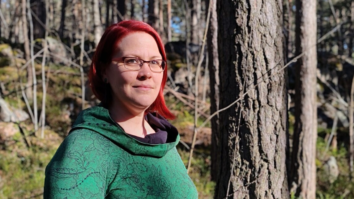 Väitöskirjatutkija Ida Penttinen, punahiuksinen nainen vihreässä mekossa katsoo kohti kameraa. Taustalla auringon valaisema metsämaasto.