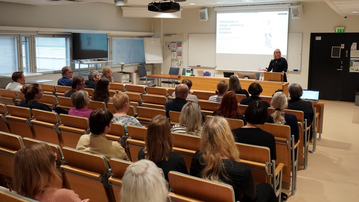 Ihmiset kuuntelevat tilan edessä puhuvaa naista.