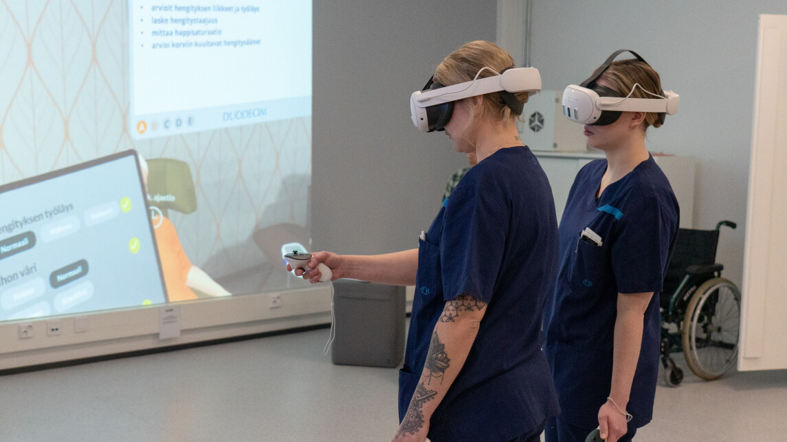 Kaksi sairaanhoitajan vaatteisiin pukeutunutta nuorta naista VR-lasit päässä. Taustalla näkyy seinälle heijastettuna VR-maailma.