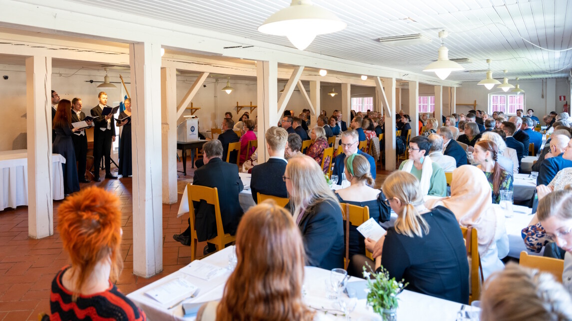 Juhlatilaisuus, jossa yleisö istuu pöytien ääressä ja katselee esiintyvää kuoroa.