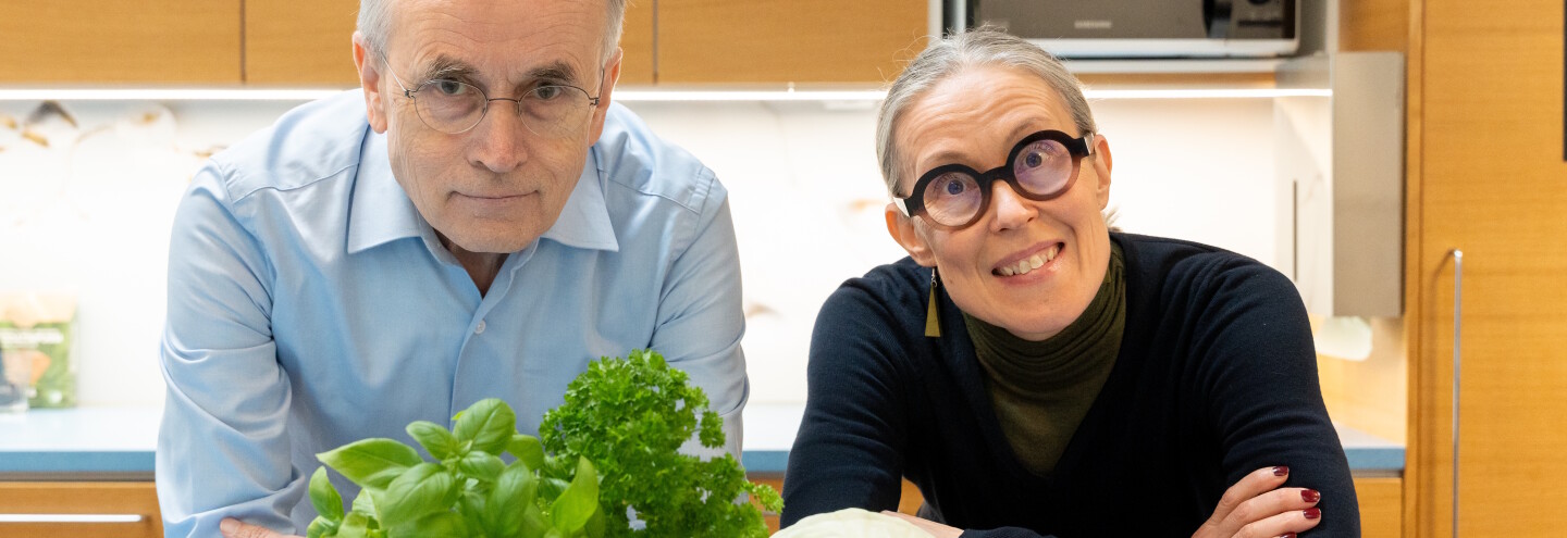 Kati Hanhineva ja Jorma Toppari nojaavat pöytään keittiössä, pöydällä vihanneksia.