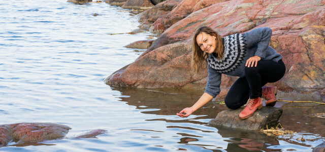 Saija Saarni rannalla kiven päällä kurottaen kädellään vettä kohti.