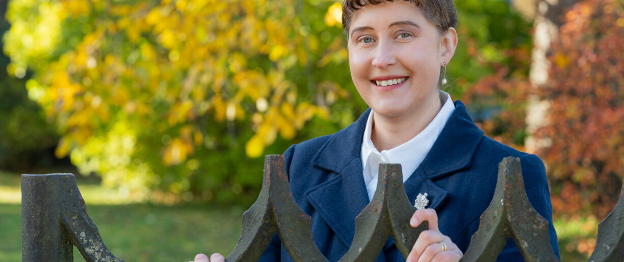 Ulla Ahlmen-Laiho lähikuvassa ulkona vanhan rautaisen portin edessä, taustalla näkyy puita joissa on värikkäät lehdet.