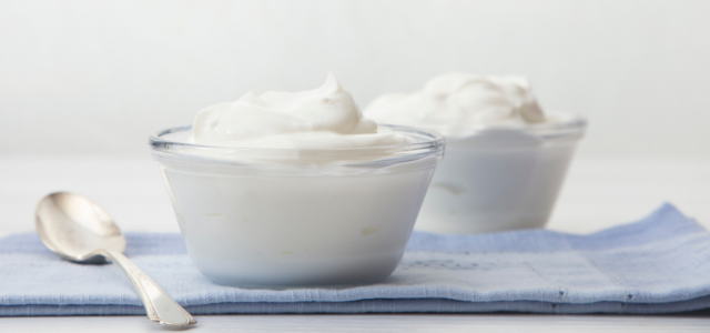 Pöydällä on lusikka ja kaksi lasikippoa, joissa on kukkuralliset annokset valkoista jogurttia.
