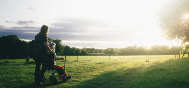 Kuvituskuvassa nuorempi henkilö työntää vanhempaa henkilöä pyörätuolissa kauniissa maisemassa