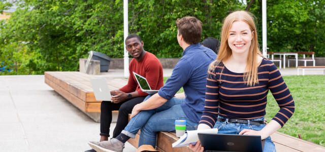 Kolme nuorta ihmistä istuu Yliopistonmäen penkeillä aurinkoisena päivänä.