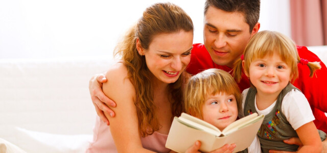 Isä, äiti ja kaksi lasta istuvat sohvalla, äiti lukee lapsille kirjaa