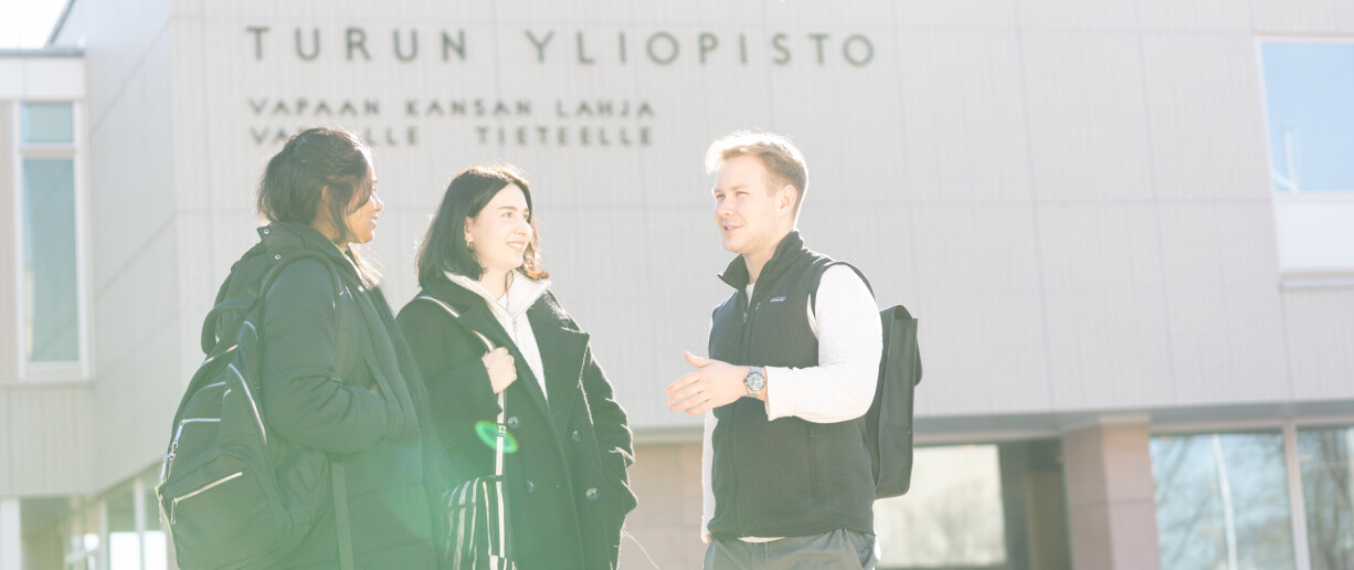 Three people chatting happily in front of the main building of the University of Turku. In the background, the wall of the university’s main building with the inscription "vapaan kansan lahja vapaalle tieteelle".