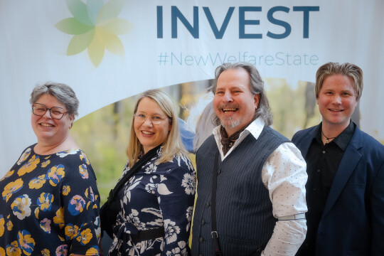 neljä ihmistä seisoo hymyilevinä INVEST-messuseinän edessä