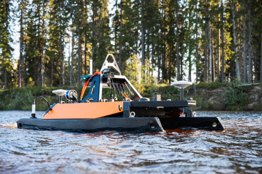 kuvassa etualalla lähikuvassa meressä oleva miehittämätön mittausalus, joka on musta-oranssi etäohjattava pikkukatamaraani, jossa on kiinni mittausvälineita ja instrumentteja. Alus on aivan lähellä rantaa ja takana näkyy mäntymetsää.