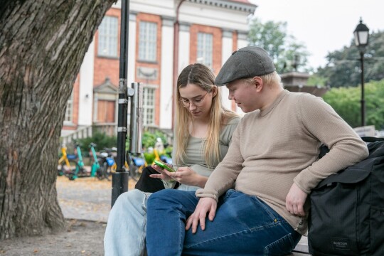 Kaksi ihmistä istuu ulkona penkillä ja katsoo puhelinta.