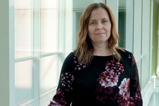 Nainen seisoo lasikäytävässä ja katsoo kameraan.