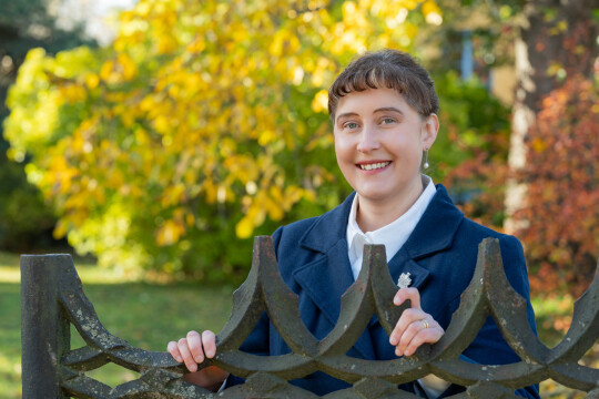 Ulla Ahlmen-Laiho lähikuvassa ulkona vanhan rautaisen portin edessä, taustalla näkyy puita joissa on värikkäät lehdet.