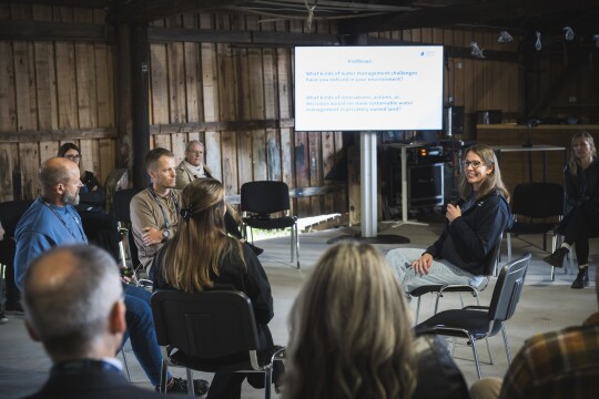 Ringissä istuvia tutkijoita keskustelemassa tilaisuudessa, kameraa kohti olevalla tutkijalla mikrofoni kädessä. Hänen takana isolla näytöllä powerpoint-esitys. Tapahtumatila saaristohenkinen, avoin iso tila jossa seinät raakalautaa ja katossa roikkuu kalastusaiheisia koristeita
