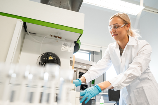 Nuori nainen työskentelee laboratoriossa yllään valkoinen labratakki, suojalasit ja kädessä siniset hanskat. Kva otettu alaviistosta ja etualalla näkyy koeputkia