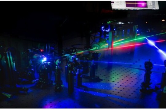 Dark laboratory setting where four different coloured lasers point to different directions, emanating a blue glow.