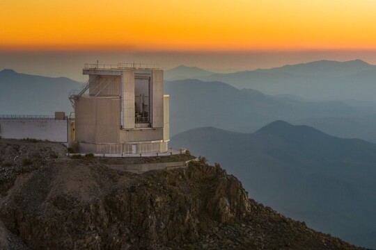 Valokuva observatioriosta vuoren rinteellä, taustalla vuoria ja oranssia taivasta.