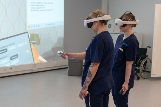 Kaksi sairaanhoitajan vaatteisiin pukeutunutta nuorta naista VR-lasit päässä. Taustalla näkyy seinälle heijastettuna VR-maailma.