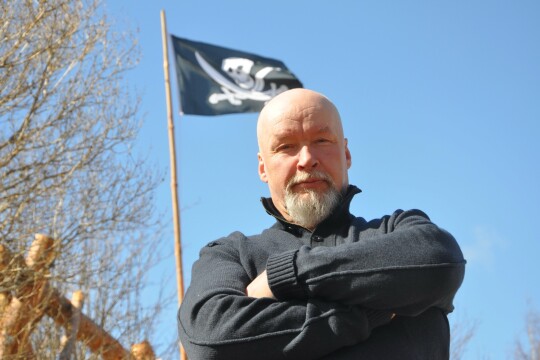 Tuomas Kuhmonen ulkona lähikuvassa, taustalla salossa merirosvolippu.