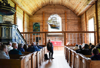Tutkija Sofia Paasikivi seisoo Seilin vanhan puukirkon penkkirivien välissä yleisön edessä ja yleisö kuuntelee kun tutkija kertoo kirkon kryptoista