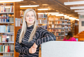 Mirva Heikkilä ketsoo kameraan ja hymyilee puolilähikuvassa, taustalla kirjahyllyjä.