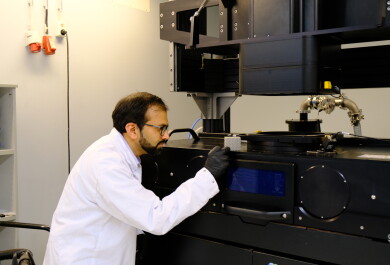 Tutkijalla on yllään valkoinen laboratoriotakki, ja hän on kumartuneena suuren mustan metallintulostuslaitteen äärelle ja tarkastelee sen päällä olevaa esinettä. Laitteen päällä, tutkijan silmänkorkeudella, on kämmenenkokoinen metallikuutio, joka on tulostettu laitteella.