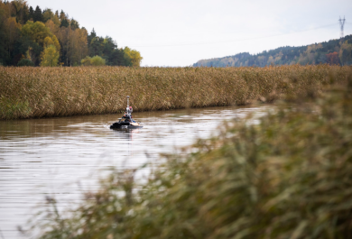 Kuvassa syksyinen rannikkomaisema, ahden kaislikon välissä olevalla kapealla vesialueella lipuu miehittämätön mittausalus, joka kerää havaintotietoa vesistöistä