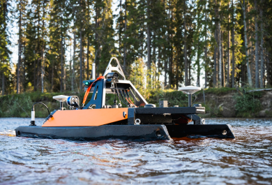kuvassa etualalla lähikuvassa meressä oleva miehittämätön mittausalus, joka on musta-oranssi etäohjattava pikkukatamaraani, jossa on kiinni mittausvälineita ja instrumentteja. Alus on aivan lähellä rantaa ja takana näkyy mäntymetsää.