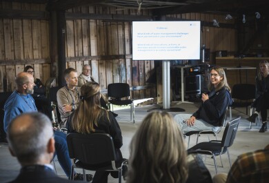 Ringissä istuvia tutkijoita keskustelemassa tilaisuudessa, kameraa kohti olevalla tutkijalla mikrofoni kädessä. Hänen takana isolla näytöllä powerpoint-esitys. Tapahtumatila saaristohenkinen, avoin iso tila jossa seinät raakalautaa ja katossa roikkuu kalastusaiheisia koristeita