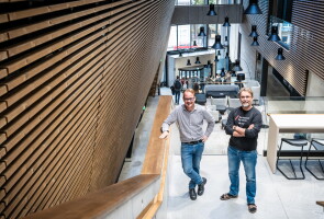 Professors Pasi Virta and Juha-Pekka Salminen are standing on the stairs in the Aurum lobby next to a wooden wall.